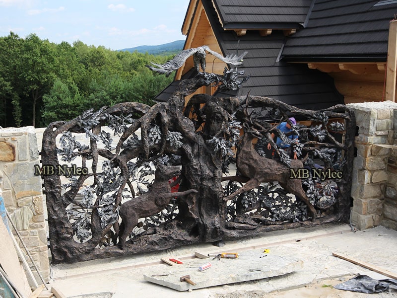 Forged driveway gate with a tree, deer and hunting dog between fieldstone piers of a log home