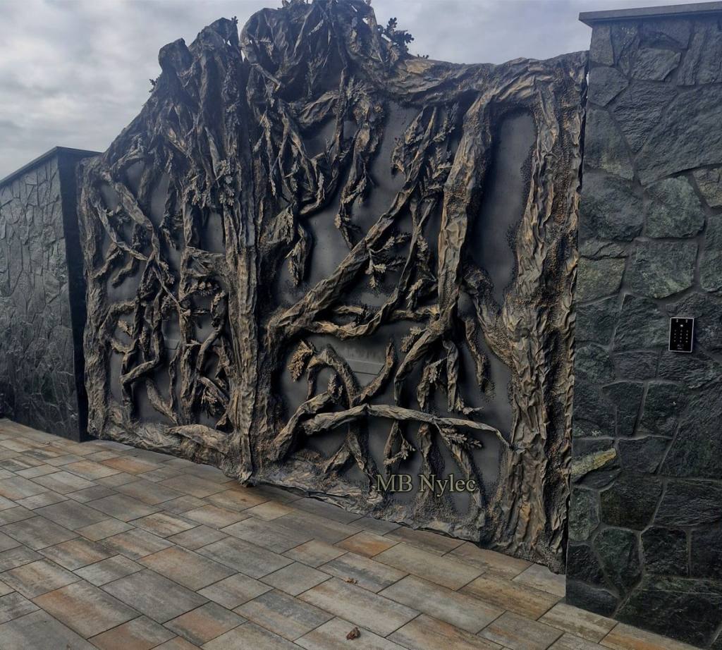 Driveway gate with a deep forged tree pattern next to a dark stone wall