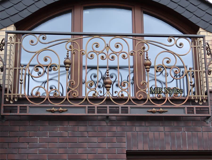 French balcony BA47 with bronze-toned scrolls and turned finials in front of an arched window on a clinker facade