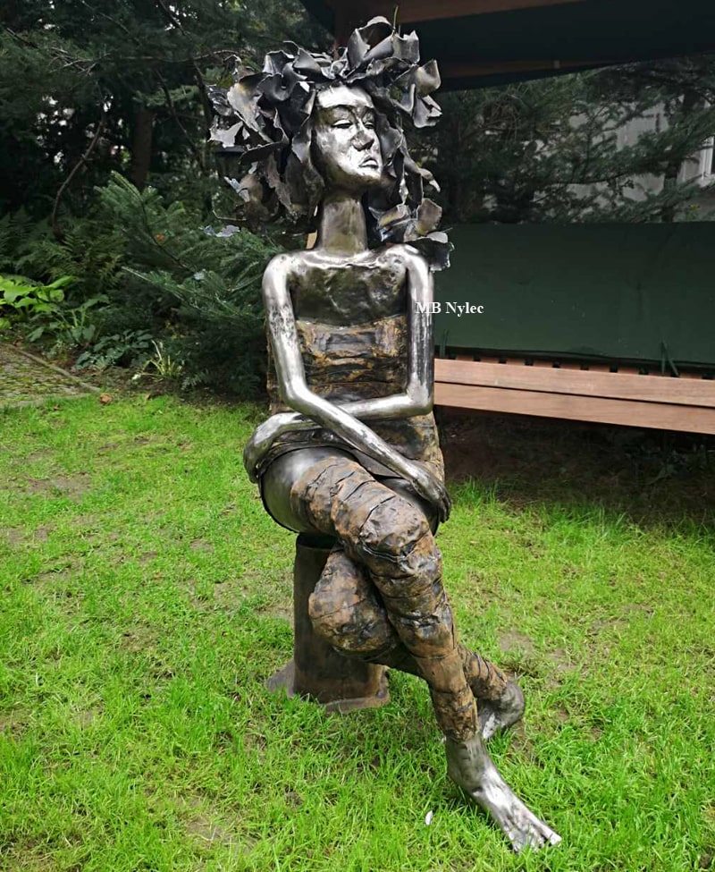 Modern steel sculpture of a woman seated by a garden bench