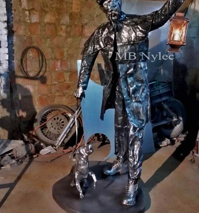 Life-size polished steel lamplighter raising a lit lantern, with a small dog on a lead
