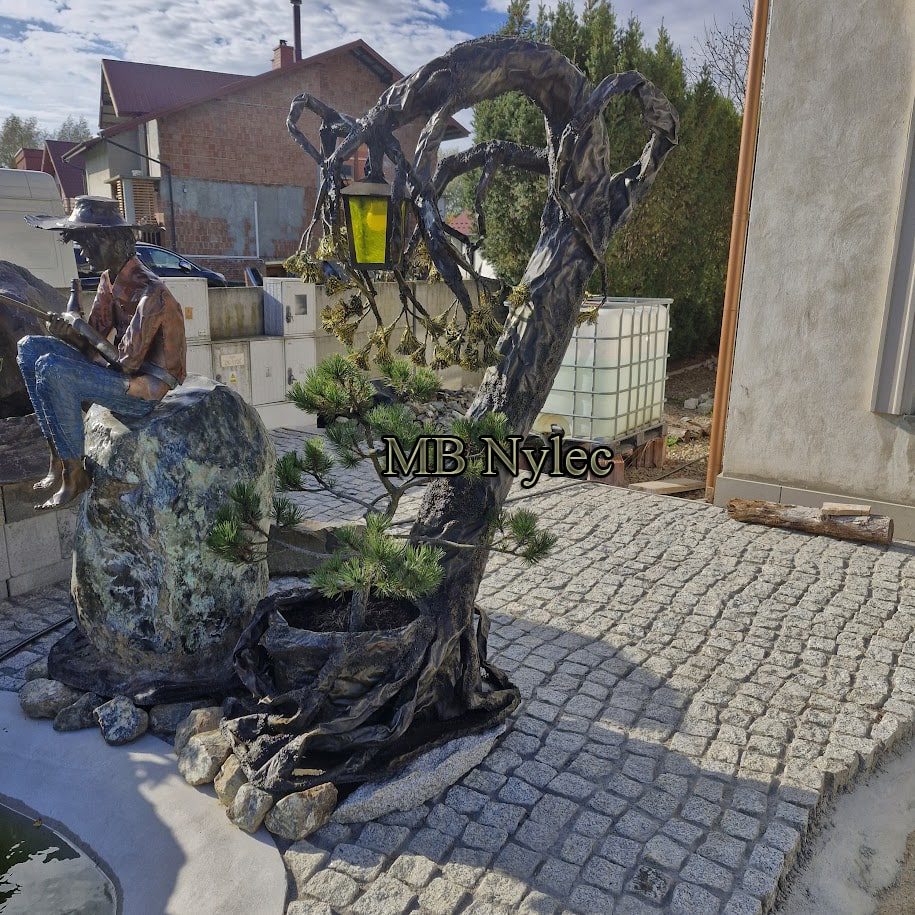 Steel tree with a lantern beside a seated figure on cobbles